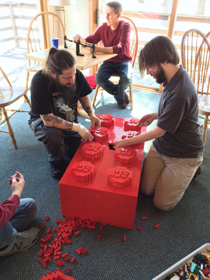 members kneeling on the floor to assemble a giant-sized red 2x4 brick - made of only 2x4 bricks